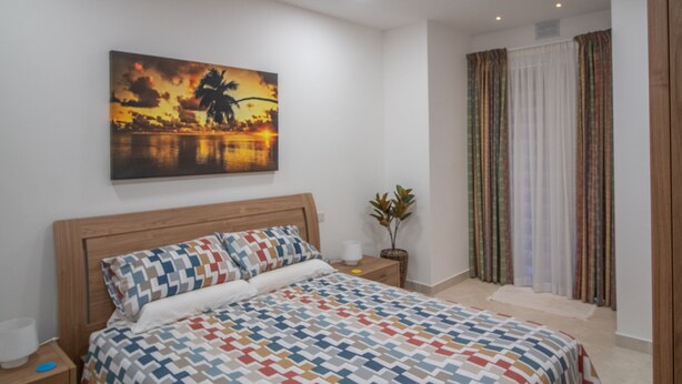 Quarto com cama, mesa de cabeceira, um quadro na parede, planta em vaso e janela com cortinas.