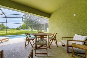 Een zwembadterras met een ronde tafel en stoelen, een comfortabele bank en uitzicht op een groen landschap door een glazen constructie.