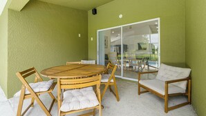 Ein überdachter Patio-Bereich mit einem runden Holztisch und Stühlen, eine Glastür, die zu einem Poolbereich führt, und eine grüne, strukturierte Wand.