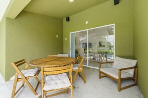 A covered patio area with a round wooden table and chairs, a glass door leading to a pool area, and a green textured wall. - Poolside Fun 8 Mi to Disney! Family Getaway (Kissimmee)