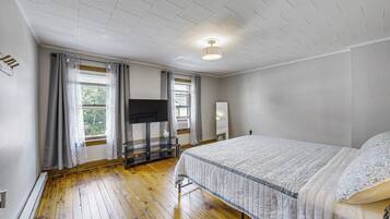 A bedroom with a wooden floor, a bed with a patterned bedspread, a flat-screen TV on a stand, and a window with curtains.