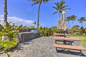 An outdoor patio area with stone flooring, a barbecue grill, picnic tables, and palm trees. - Halii Kai Condo Close in Proximity to Ocean/Pool (Waikoloa Village)
