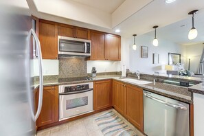 A modern kitchen with wooden cabinets, stainless steel appliances, granite countertops, and pendant lighting. - Halii Kai Condo Close in Proximity to Ocean/Pool (Waikoloa Village)
