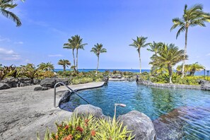 A clear blue pool with a stone deck, surrounded by palm trees and rocks, overlooking the ocean. - Halii Kai Condo Close in Proximity to Ocean/Pool (Waikoloa Village)