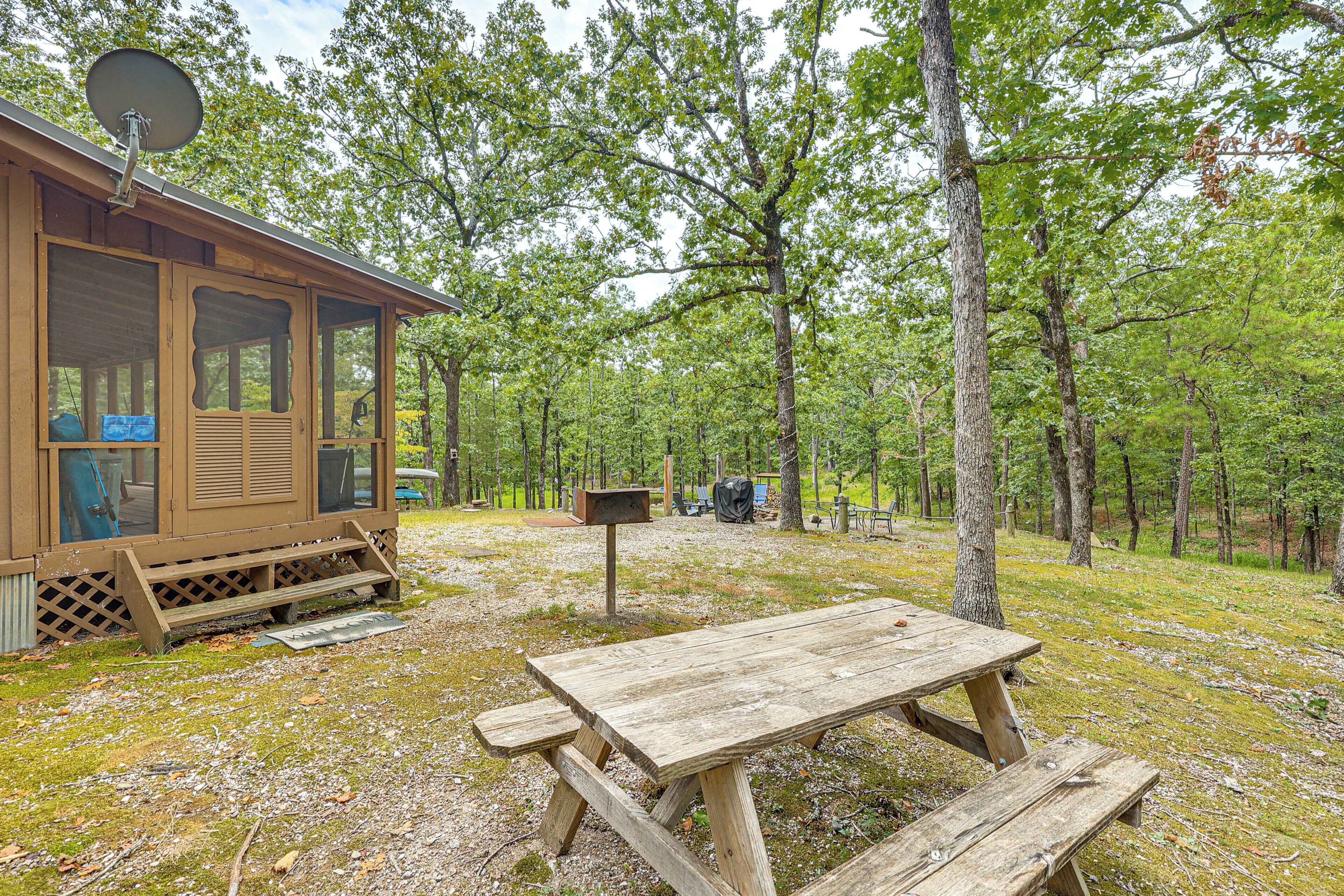 A wooden cabin with a satellite dish, a picnic table, and a mailbox in a forested area.