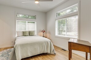 Une chambre à coucher avec un lit, deux fenêtres équipées de stores, une table de chevet, une lampe et une table en bois.