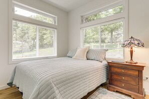 Ein Schlafzimmer mit einem Bett, einem Nachttisch und einer Lampe. Große Fenster mit Jalousien sind vorhanden.