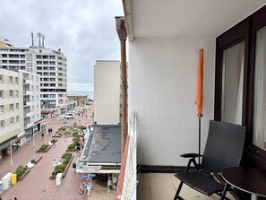 Un balcón con una silla y una mesa, con vista a una calle peatonal con tiendas y un edificio de techo plano.