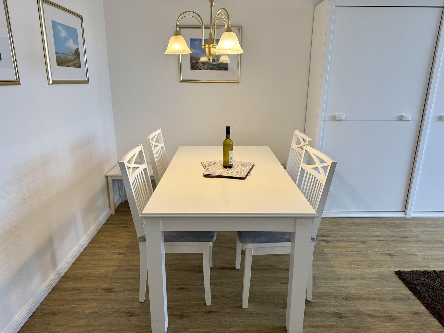 A dining area with a white table, four chairs, a chandelier, and framed pictures on the wall.
