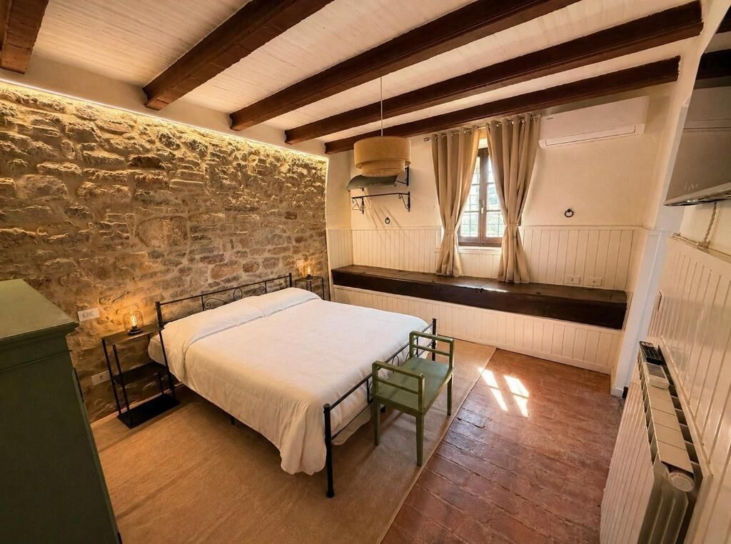 A bedroom with a stone wall, a bed, a chair, a window with curtains, and a radiator.