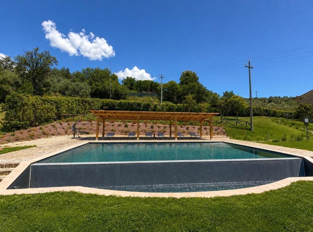 A rectangular swimming pool with a wooden pergola and lounge chairs, surrounded by a landscaped garden with a grassy area and a hilly backdrop.
