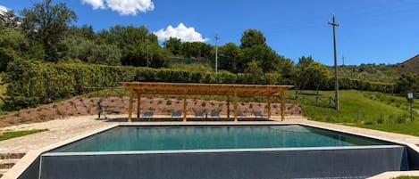 A rectangular swimming pool with a wooden pergola and lounge chairs, surrounded by a landscaped garden with a grassy area and a hilly backdrop.