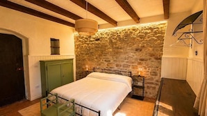 Un dormitorio con pared de piedra, una cama, una silla verde, un banco de madera y un aplique colgante.