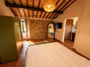 A bedroom with a bed, a stone wall, a television, and a kitchen area.
