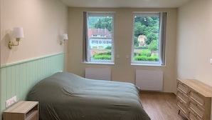 Quarto com cama, penteadeira, duas janelas com cortinas e luminárias fixadas na parede.