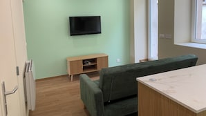 Quarto com televisão de tela plana na parede, um armário de madeira abaixo, sofá verde, mesa com tampo de mármore e janela com vista.