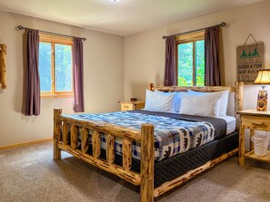 Ein Zimmer mit einem hölzernen Bett, zwei mit Vorhängen versehenen Fenstern, einem Nachttisch und einer Wanddekoration.