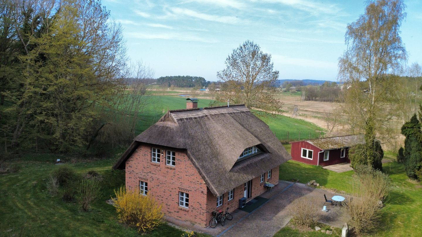Een huis met rieten dak en bakstenen gevel, omgeven door een groen landschap en bomen.