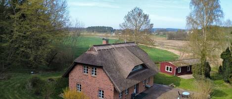 Een huis met rieten dak en bakstenen gevel, omgeven door een groen landschap en bomen.