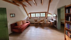 Een kamer met een schuine plafond, een rode bank, twee stoelen, een bureau en een boekenkast.