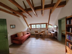 Ein Zimmer mit schrägem Dach, einem roten Sofa, zwei Stühlen, einem Schreibtisch und einem Bücherregal.