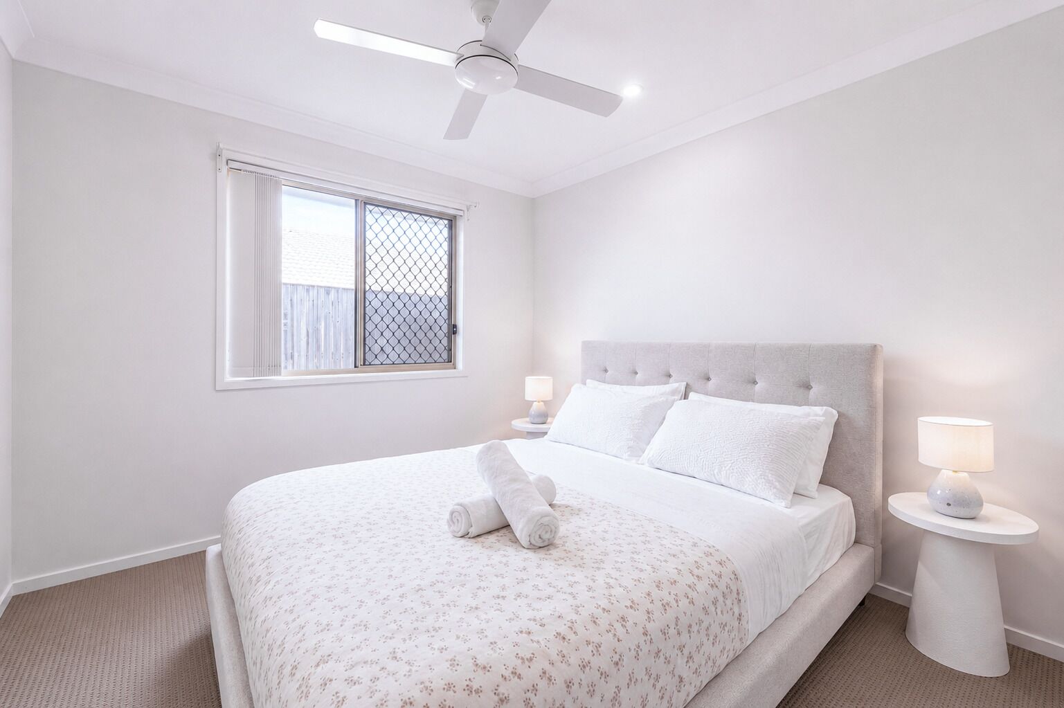 A bedroom with a bed, a ceiling fan, a window with a metal screen, and two bedside lamps.