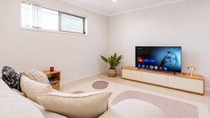 A modern living room with a flat-screen TV, a sofa, a coffee table, a plant, and a window with blinds. - The Garden Nook | Duplex w/ Private Backyard (Waterford West)