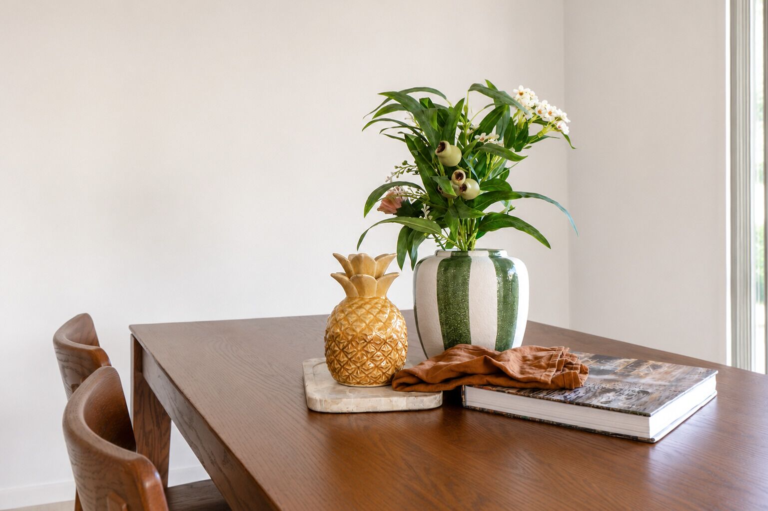 Un tavolo di legno con un vaso a forma di ananas, un vaso a righe con fiori e un libro.