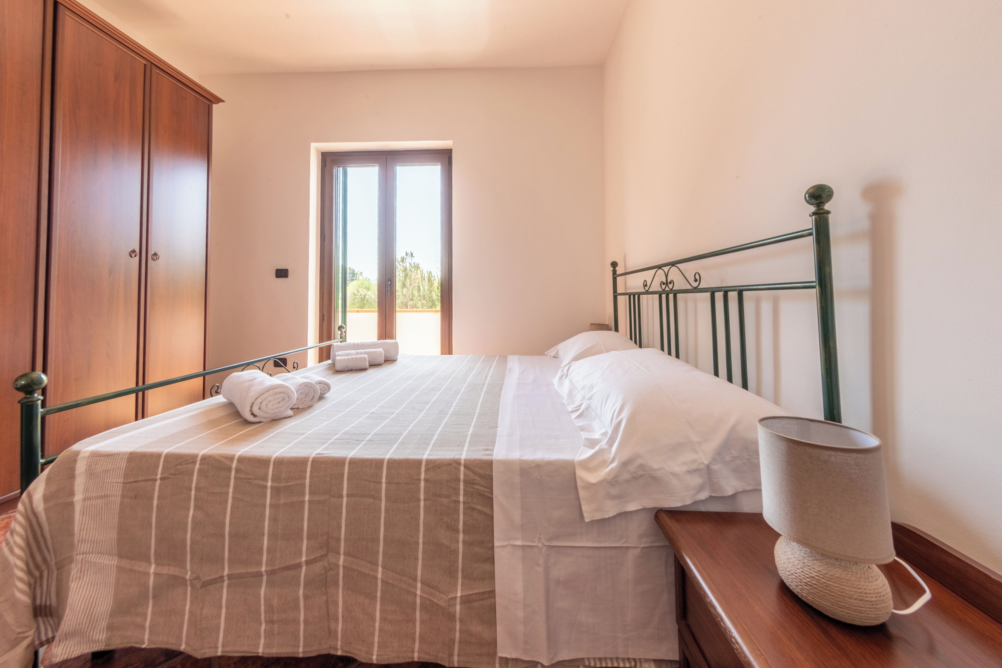 A bedroom with a bed, a wooden wardrobe, a window, and a bedside lamp.
