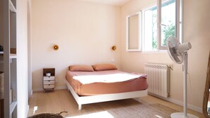 Une chambre à coucher avec un lit, un ventilateur sur pied, un radiateur et une fenêtre donnant sur des arbres.