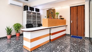 Un área de recepción con piso de mármol, un mostrador de recepción, dos plantas en macetas, un ventilador de techo y una puerta de madera.
