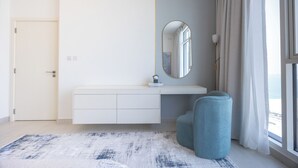 A modern bedroom with a white vanity, a round mirror, a blue chair, and a view of the sea through large windows. - Bloomfields 2BR in The Bridges - Reem (Abu Dhabi)
