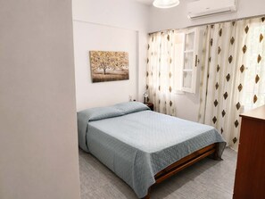 A bedroom with a bed, a bedside table, a lamp, a window with curtains, and an air conditioning unit. - Kalymnos Island Pearl - Seaside Getaway (Kalymnos)