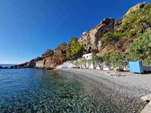 Ein Kiesstrand mit klarem Wasser, eine felsige Klippe und ein Gebäude mit weißer Fassade.
