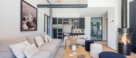 Un salon moderne comprenant un canapé, une table basse et un pouf, une cheminée et donnant sur la salle à manger.