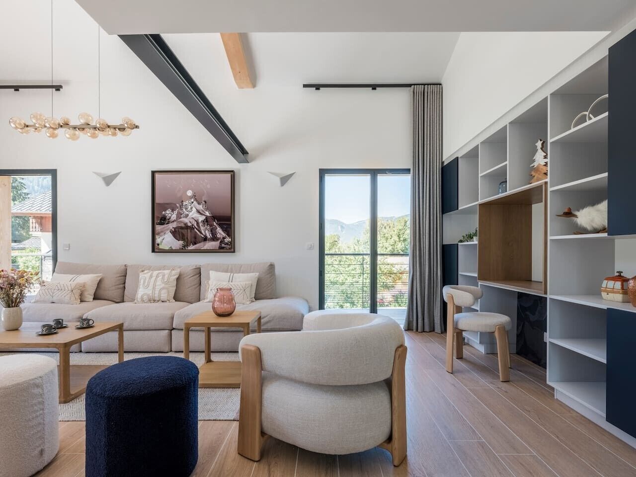 A modern living room with a sofa, coffee table, and armchairs, a bookshelf, and a large window with a view.