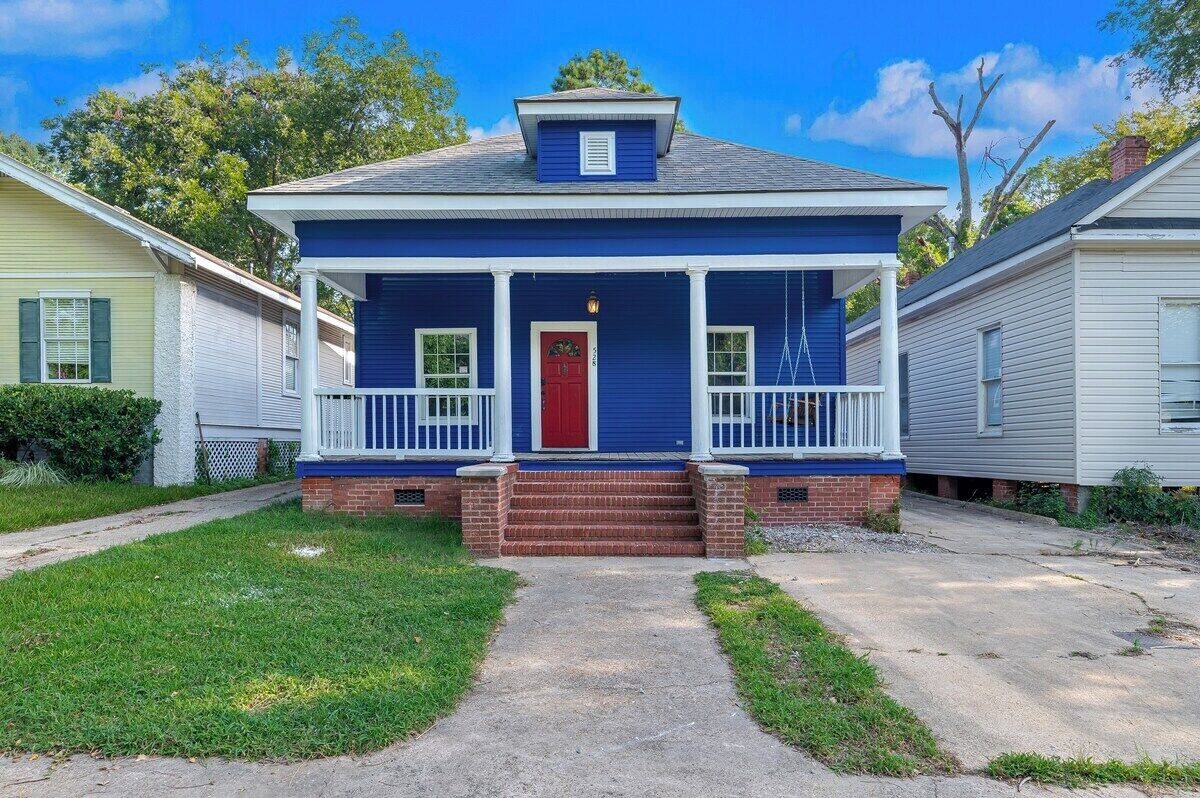 A two-story house with a red door, a front porch with white railings, and a brick foundation.