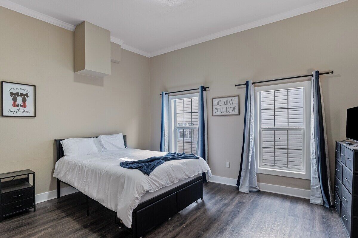 Un dormitorio con cama, mesita de noche, cómoda y dos ventanas con cortinas.