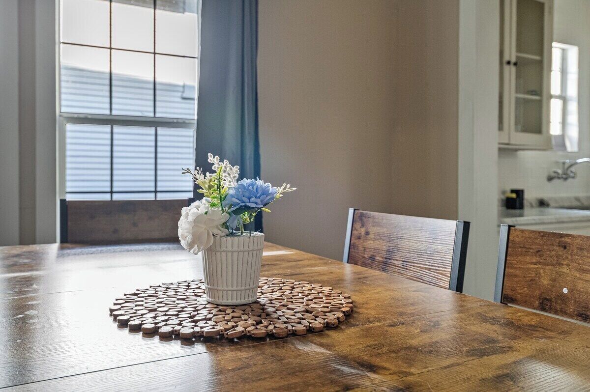 Een houten eettafel met een witte vaas met bloemen, een geweven placemat en een raam met jaloezieën op de achtergrond.