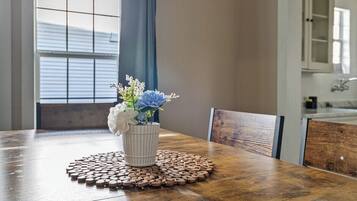 Een houten eettafel met een witte vaas met bloemen, een geweven placemat en een raam met jaloezieën op de achtergrond.