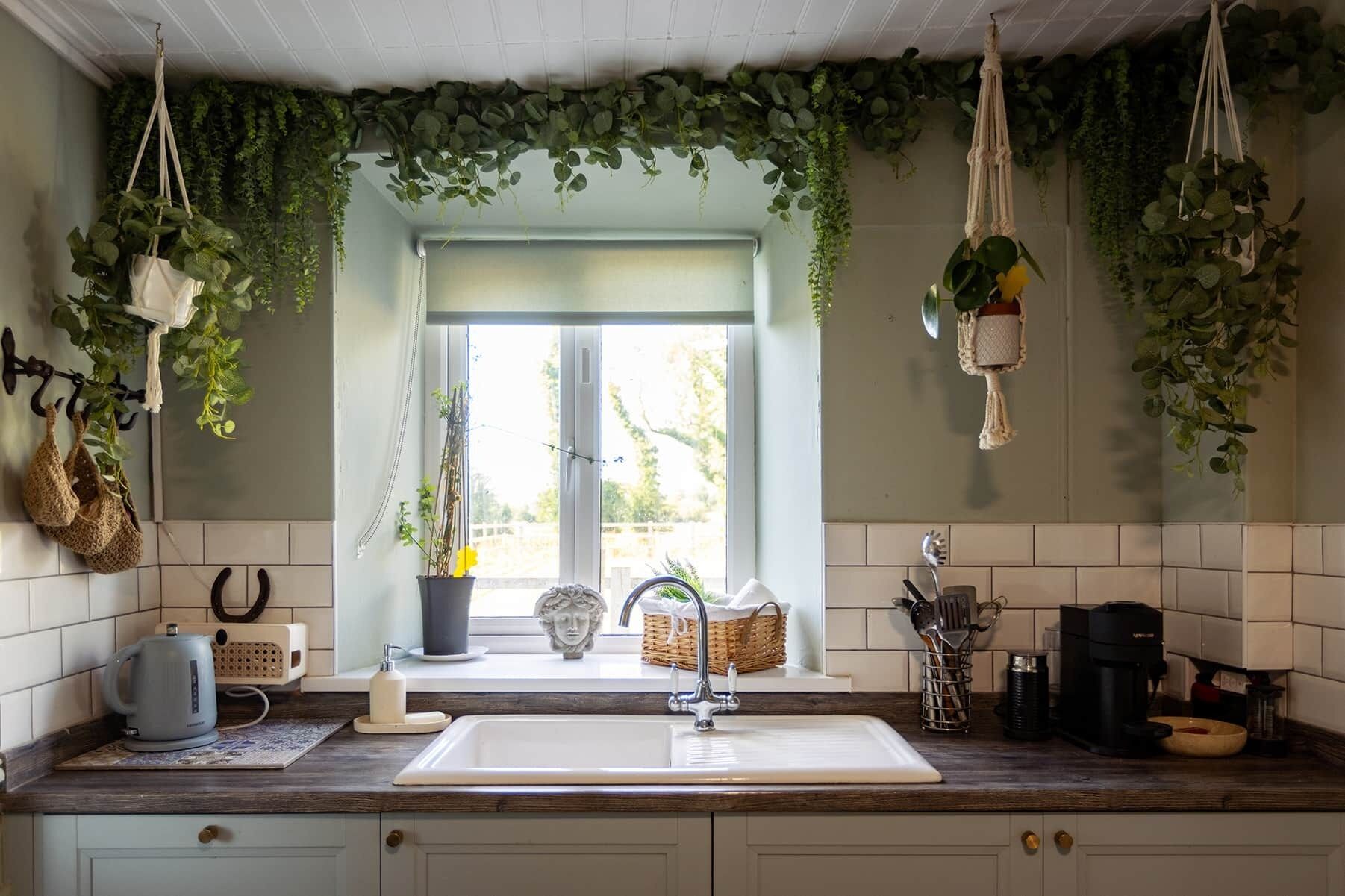 Eine Küche mit einem weißen Spülbecken, einem Wasserkocher und einer Kaffeemaschine. Hängende Pflanzen und ein Fenster mit Blick.