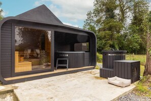 A modern, black wooden cabin with a large sliding glass door, a small outdoor seating area, and a hot tub. - Bealtaine (Síor Collection) (Carrick-On-Shannon)