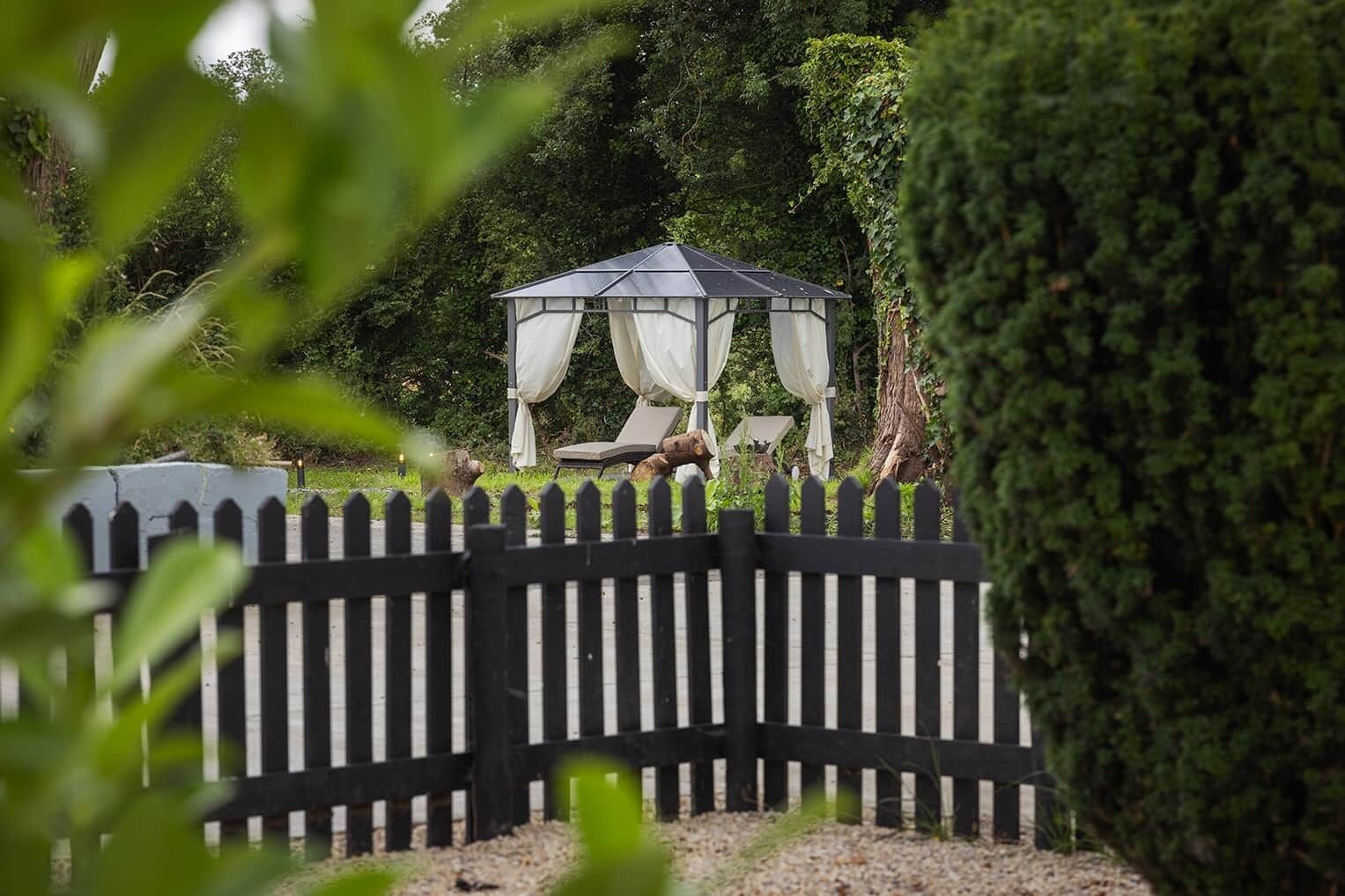 A gazebo with a roof and white curtains, surrounded by a black picket fence, set in a garden with dense foliage.