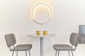 Una mesita redonda con dos sillas, dos tazas amarillas y una lámpara circular fijada a la pared.