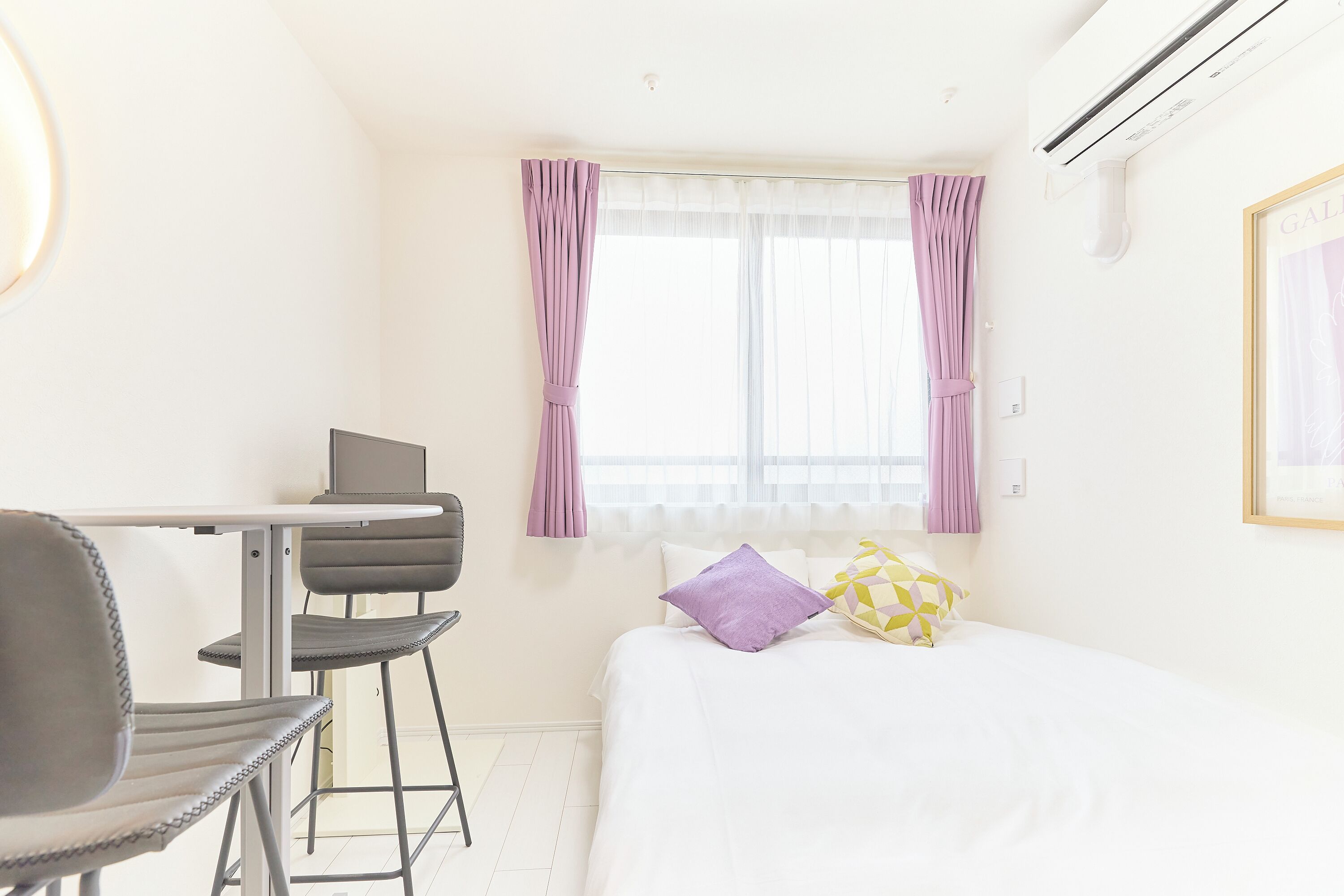A hotel room with a bed, a desk with a chair, a window with curtains, and an air conditioning unit.