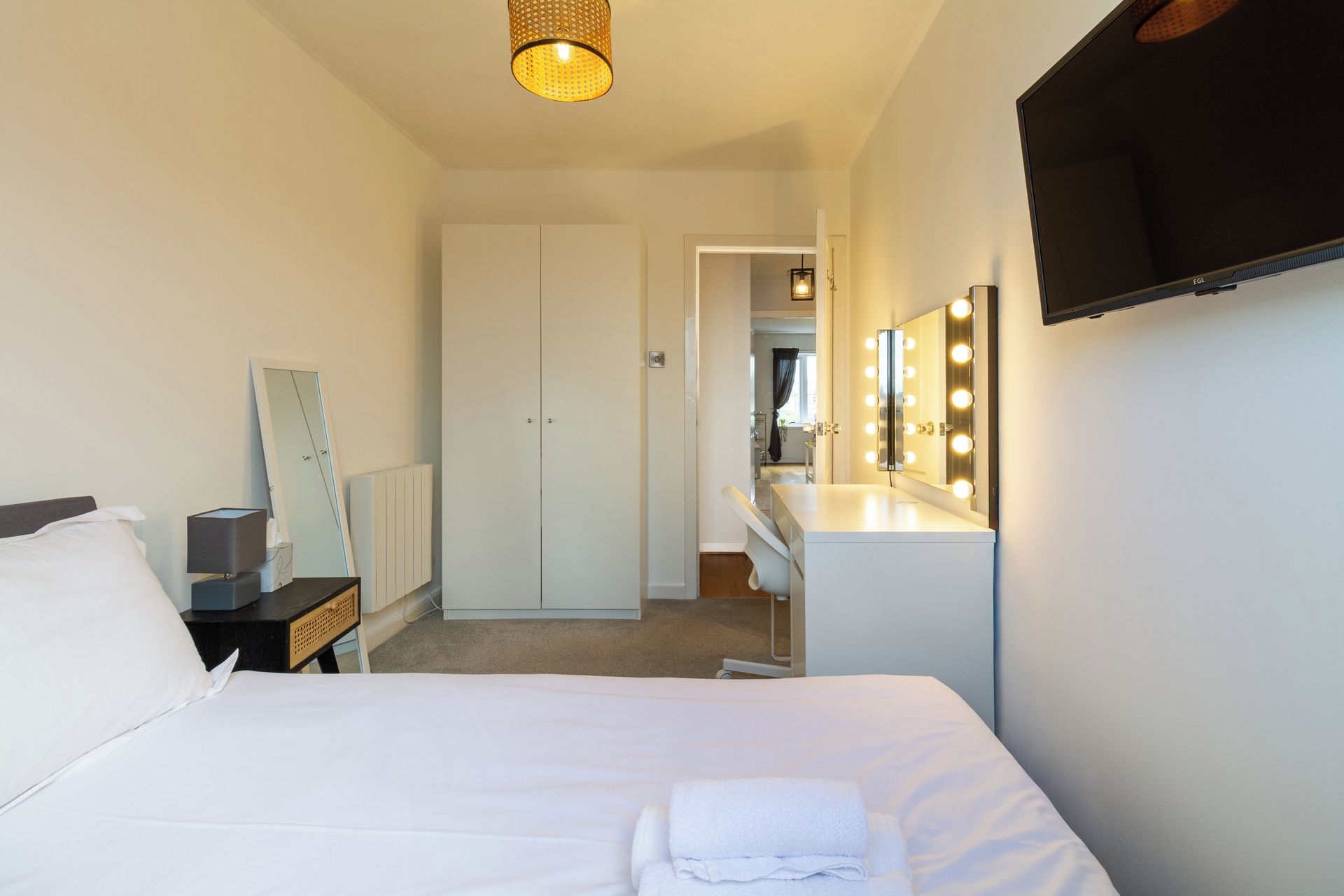 A hotel room with a bed, a bedside table with a lamp, a dresser, a mirror with lights, and a wardrobe.