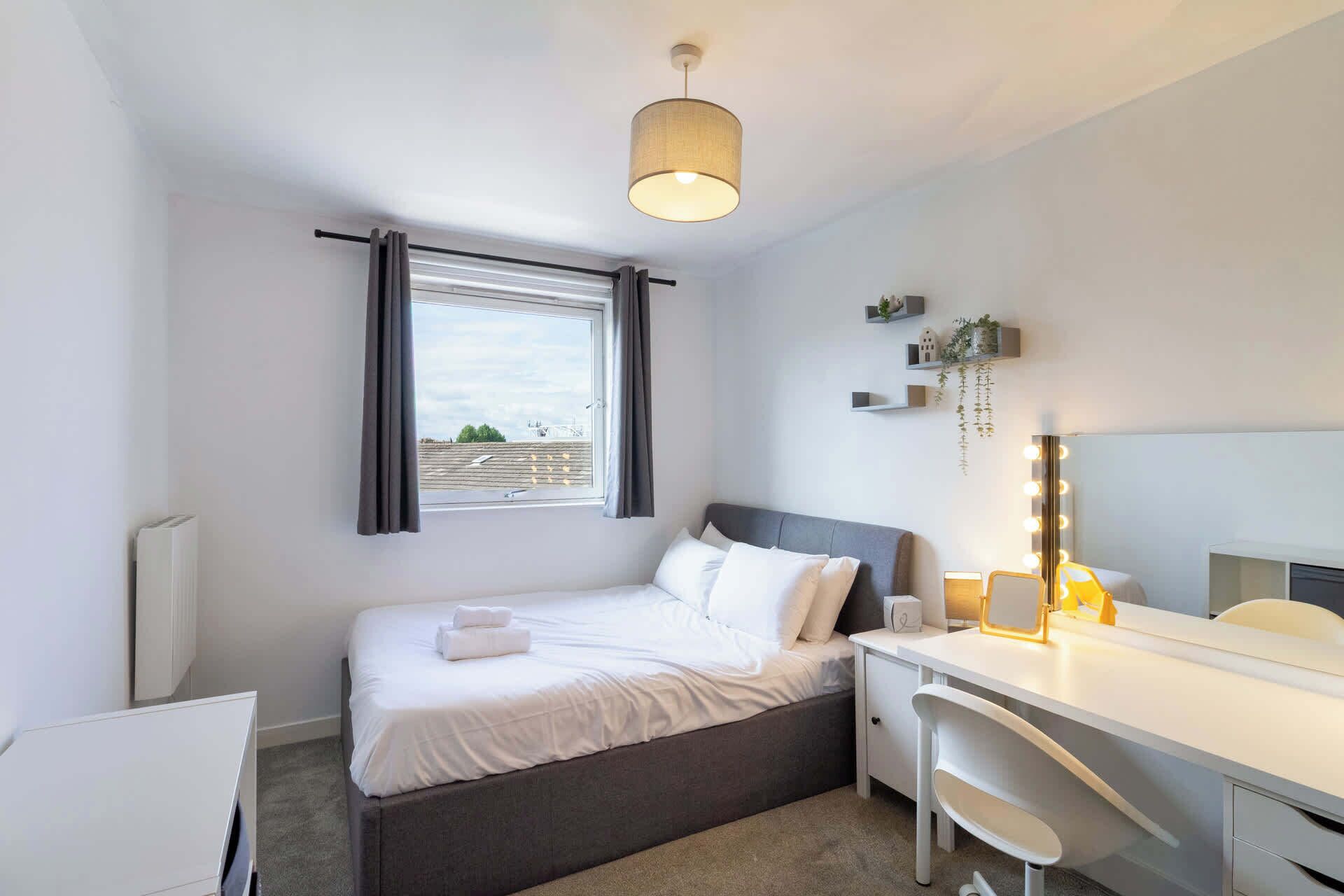 A bedroom with a bed, a desk, a mirror with lights, a window with curtains, and a radiator.