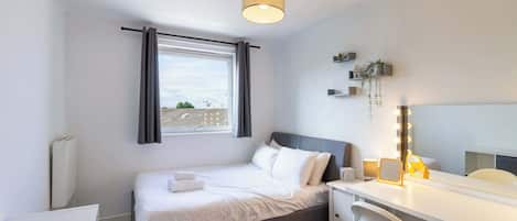 A bedroom with a bed, a desk, a mirror with lights, a window with curtains, and a radiator.