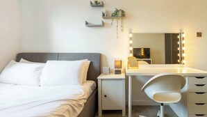 Quarto com cama, mesa de cabeceira, escrivaninha com cadeira, espelho com iluminação e prateleiras na parede.