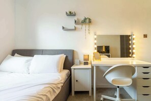 Ein Schlafzimmer mit Bett, Nachttisch, Schreibtisch mit Stuhl, Spiegel mit Beleuchtung und Regalen an der Wand.
